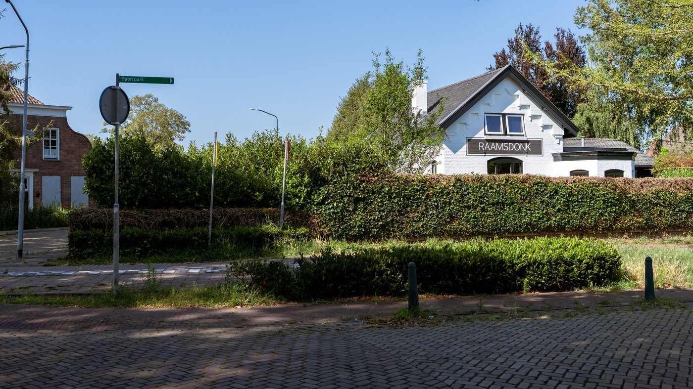 Hetzelfde pand nu, wit gepleisterd achter een haag, met het bord RAAMSDONK nog op de gevel.